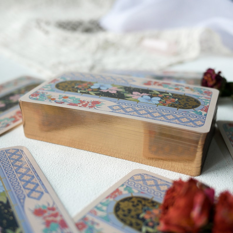 May include: A deck of tarot cards with a gold gilded edge and a colorful floral design on the back. The cards are stacked on a white surface.