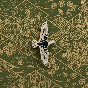 Pode incluir: Um broche de esmalte preto e branco de um pássaro em voo contra um fundo estampado verde e dourado.