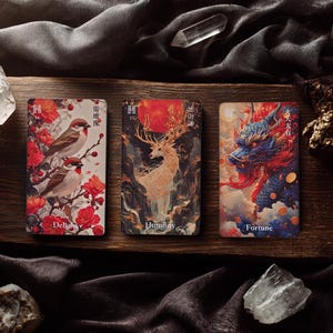 May include: Three rectangular oracle cards featuring artistic illustrations. The left card shows two birds among red flowers with the text Delight. The centre card depicts a deer with large antlers and a red sun labelled Humility. The right card shows a blue dragon with the text Fortune.