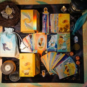 May include: An assortment of tarot cards and related items, including a deck box, drawstring bag, and decorative elements. The cards feature colorful illustrations with the text "luno with the sun". The color palette includes yellow, blue, and orange.