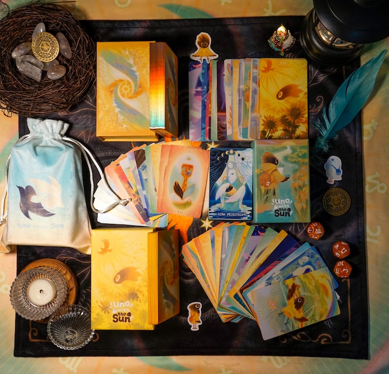 May include: An assortment of tarot cards and related items, including a deck box, drawstring bag, and decorative elements. The cards feature colorful illustrations with the text "luno with the sun". The color palette includes yellow, blue, and orange.