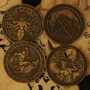 May include: Four round, bronze-colored metal tokens with intricate designs. Each token features a different image: a woman with multiple arms, a creature with wings, a figure with a raised arm, and a horned figure with wings.