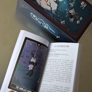 May include: An open tarot card deck featuring a card titled "The Hanged Man." The card depicts a cartoon figure hanging upside down. The box has a dark blue background with a skeleton and other cartoon figures.