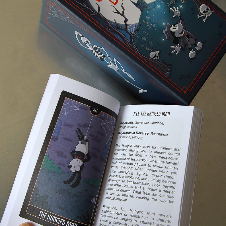 May include: An open tarot card deck featuring a card titled "The Hanged Man." The card depicts a cartoon figure hanging upside down. The box has a dark blue background with a skeleton and other cartoon figures.