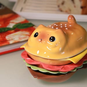May include: A novelty ceramic cat-shaped burger. The top bun is yellow-brown with white dots, the cheese is yellow, and the lettuce and tomato are green and red. The burger is on a reflective surface.