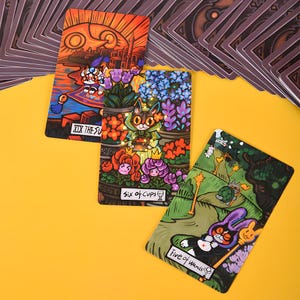 May include: Three tarot cards with colorful illustrations are displayed on a yellow surface. The cards feature whimsical art with text including "XIX The Sun", "Six of Cups", and "Five of Wands". A deck of cards is visible in the background.