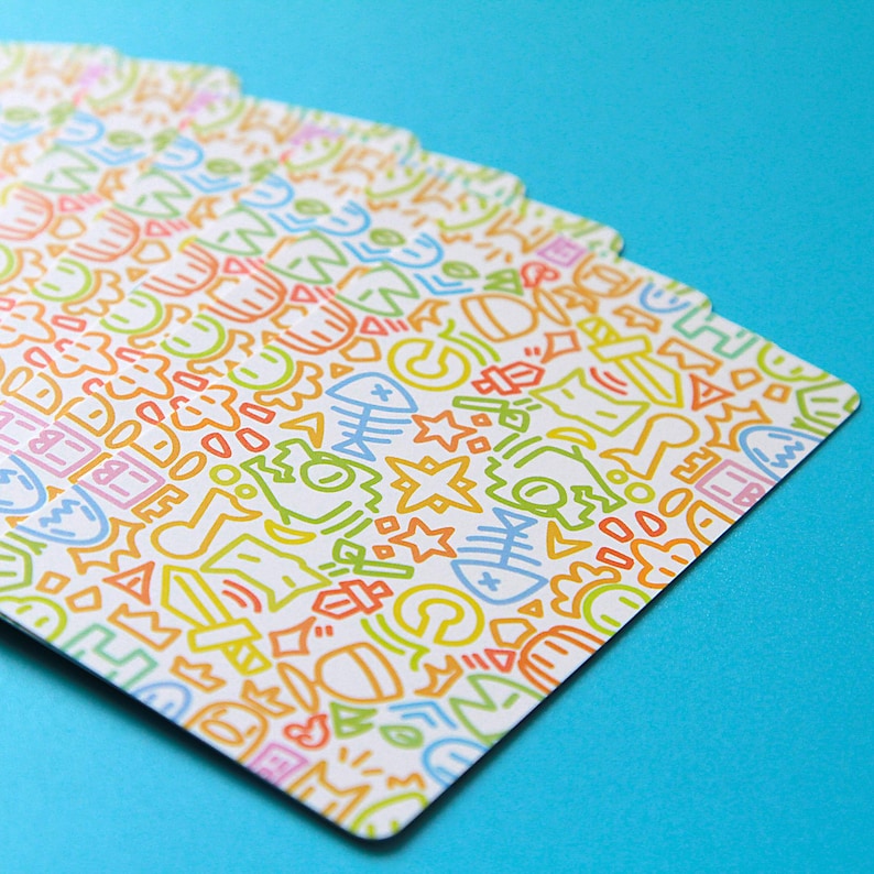 May include: A fan of playing cards with a white background and a colorful doodle pattern. The doodles include fish bones, stars, and other abstract shapes in orange, green, blue, and pink. The cards are fanned out against a bright blue background.