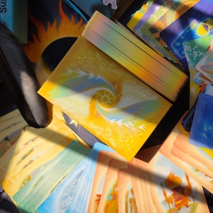 May include: A deck of oracle cards with a yellow and blue spiral design, titled "Calling the Sun." The cards are surrounded by other cards with colorful illustrations, a black feather, and a box with the text "Calling the Sun."