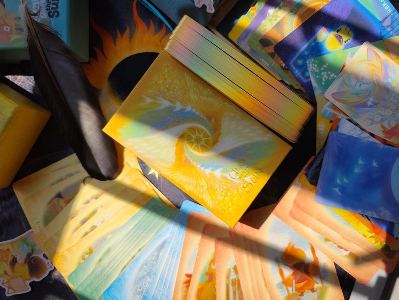 May include: A deck of oracle cards with a yellow and blue spiral design, titled "Calling the Sun." The cards are surrounded by other cards with colorful illustrations, a black feather, and a box with the text "Calling the Sun."
