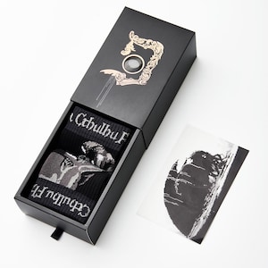 Puede incluir: Una caja negra con un diseño dorado en la parte superior. La caja está abierta y contiene un par de calcetines negros con un estampado gráfico blanco de una criatura. El texto "Cthulhu" está impreso en los calcetines. Una foto en blanco y negro de un paisaje está al lado de la caja.