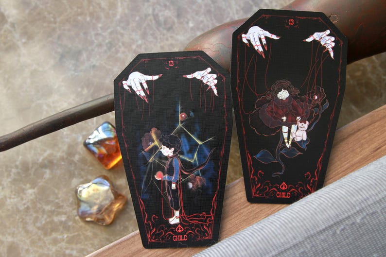 May include: Two coffin-shaped cards with black backgrounds and red accents. Each card features the number 13, the word CHILD, and an illustration of a child being controlled by puppet strings held by white hands with red fingernails.