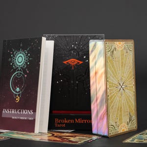 May include: A deck of Broken Mirror Tarot cards with a gold-edged box, a black box featuring an orange eye graphic, and an instruction booklet with a teal circular design. Several individual tarot cards are spread out in the foreground.
