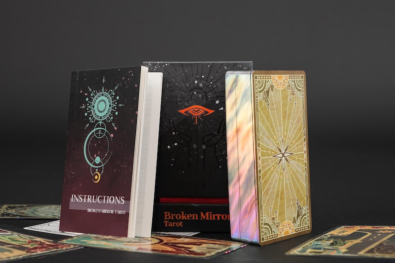 May include: A deck of Broken Mirror Tarot cards with a gold-edged box, a black box featuring an orange eye graphic, and an instruction booklet with a teal circular design. Several individual tarot cards are spread out in the foreground.