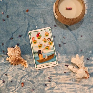 May include: A tarot card reading with a "Seven of Cups" card, a seashell, a coconut candle, and decorative stones on a blue tie-dye cloth. The card depicts a woman in a boat with a cat, surrounded by cups with various images.
