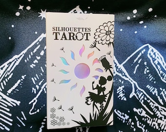 May include: A white rectangular tarot deck box titled Silhouettes Tarot in black text. The design features a central iridescent sun, a black silhouette of a figure standing near a large flower, scattered dandelion seeds, and small floral accents.