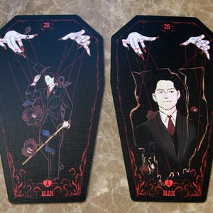 May include: Two coffin-shaped cards featuring a black background with red accents. Each card displays the number 28 at the top, the letter A at the bottom, and the word MAN. The illustrations depict a figure in a suit controlled by marionette strings.