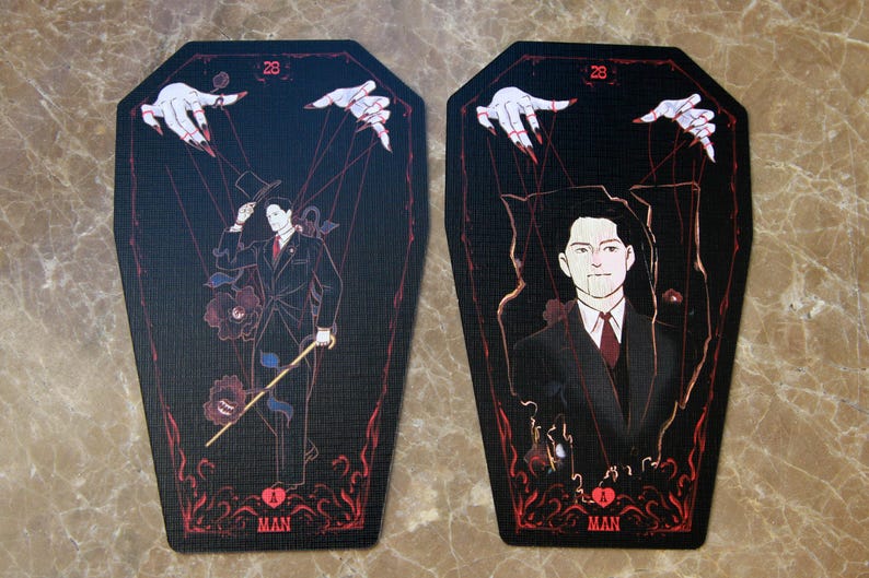 May include: Two coffin-shaped cards featuring a black background with red accents. Each card displays the number 28 at the top, the letter A at the bottom, and the word MAN. The illustrations depict a figure in a suit controlled by marionette strings.