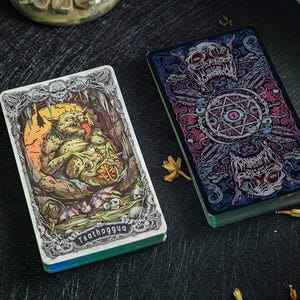 May include: Two rectangular playing cards with intricate illustrations. The left card features a green, toad-like creature labeled Tsathoggua in a cave setting. The right card displays a dark, symmetrical design with skulls and a central eye motif.