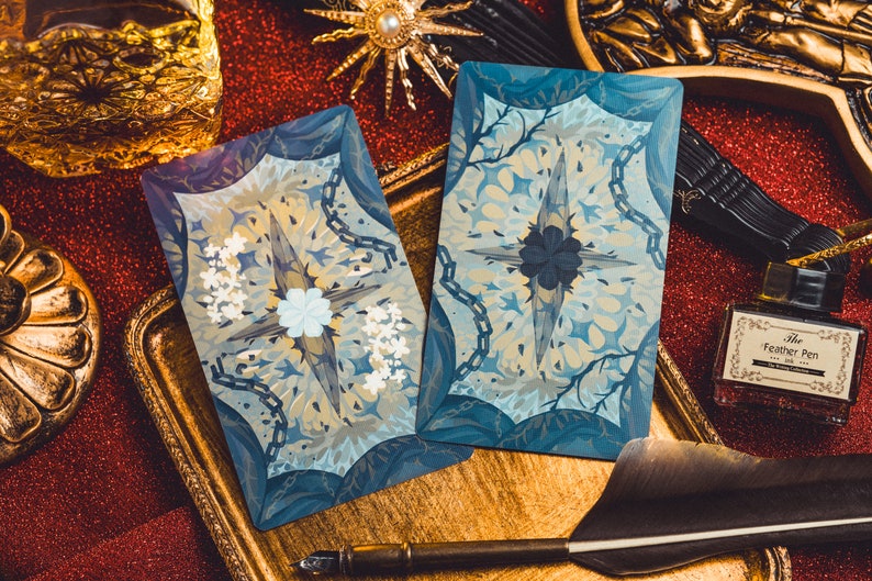 Op de afbeelding: Twee tarotkaarten met een blauw en wit bloemenontwerp, met een centraal sterrenpatroon. De kaarten liggen op een gouden dienblad, samen met een ganzenveer en een inktfles met het opschrift "The Feather Pen".