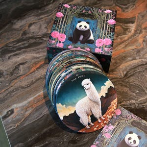 May include: A deck of round tarot cards featuring illustrations of animals and nature scenes. The cards are stacked next to a box with a panda illustration. The card on top features an alpaca. The box and cards have a floral design.