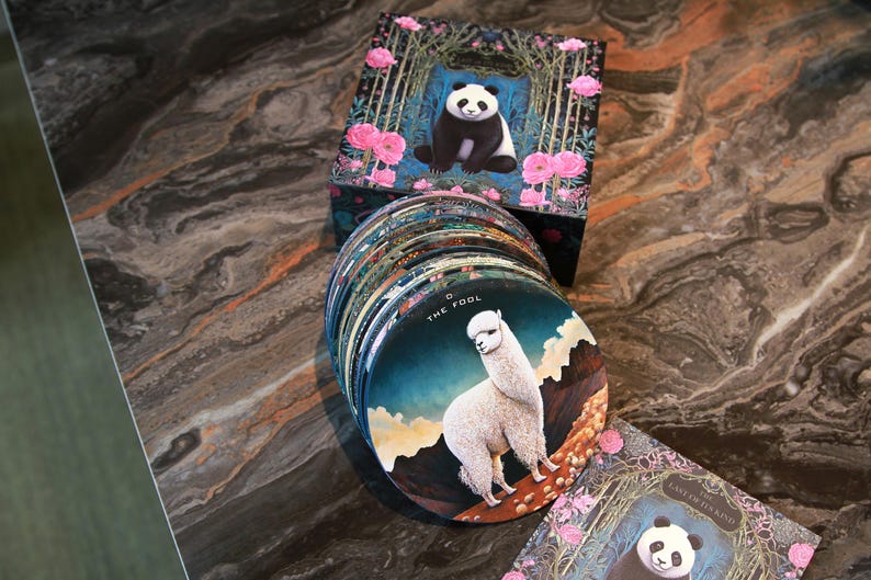 May include: A deck of round tarot cards featuring illustrations of animals and nature scenes. The cards are stacked next to a box with a panda illustration. The card on top features an alpaca. The box and cards have a floral design.