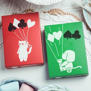 Puede incluir: Dos barajas de cartas, una roja y otra verde, con ilustraciones caprichosas. La baraja roja presenta un gato blanco sosteniendo globos con forma de símbolos de cartas. La baraja verde muestra un perro con un cañón de confeti con forma de símbolos. Ambas barajas están sobre una superficie blanca.
