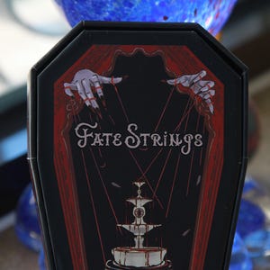 May include: A black coffin-shaped box featuring a red and black graphic illustration. The design depicts two pale hands with long dark fingernails manipulating red strings above a fountain. The text Fate Strings is printed in white.