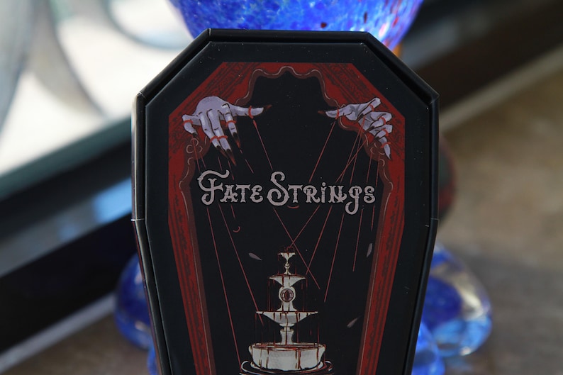 May include: A black coffin-shaped box featuring a red and black graphic illustration. The design depicts two pale hands with long dark fingernails manipulating red strings above a fountain. The text Fate Strings is printed in white.