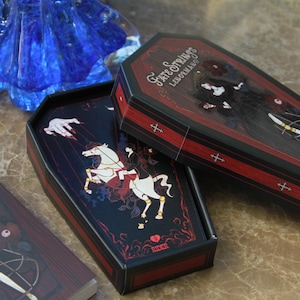 May include: A coffin-shaped deck of Fate Strings Lenormand cards featuring black and red illustrations. The box lid displays a figure in black, while the open box reveals a card with a white horse and the text Rider. A separate card shows the text Fate Strings Lenormand.