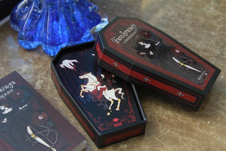 May include: A coffin-shaped deck of Fate Strings Lenormand cards featuring black and red illustrations. The box lid displays a figure in black, while the open box reveals a card with a white horse and the text Rider. A separate card shows the text Fate Strings Lenormand.