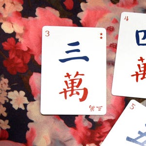 Pu&ograve; includere: Primo piano di tessere Mahjong bianche con caratteri cinesi e numeri blu e rossi. La tessera centrale mostra il numero 3, con i caratteri di "tre" e "miriade" in blu e rosso. Lo sfondo presenta un motivo floreale.