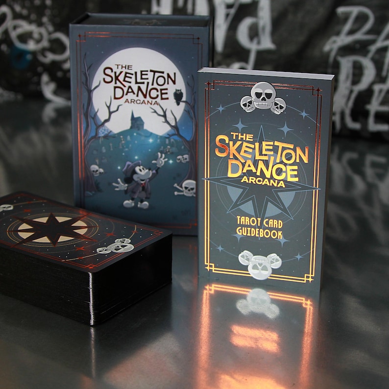 May include: A tarot card set titled "The Skeleton Dance Arcana." The set includes a box, a guidebook, and a deck of cards. The box and guidebook feature illustrations of a cartoon character, skulls, and a full moon. The cards have a black background with a star design.
