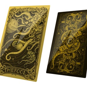 May include: Two rectangular cards with gold and black ink illustrations. The left card features a tentacled creature, planets, and the text THE FOOL. The right card displays a complex, symmetrical design with a central eye and skull motifs.