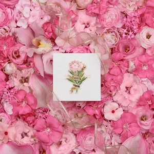 May include: A gold-toned enamel pin featuring a pink flower bouquet with green leaves and a gold banner. The pin is surrounded by pink flowers.