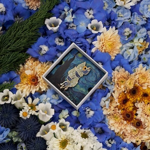 May include: A gold and blue enamel pin featuring a stylized bird with a single eye, set against a background of a starry night sky. The pin is displayed in a white box with a blue and white background.