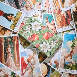 May include: A green tarot card box with a floral and pomegranate design, surrounded by tarot cards. The cards feature illustrations of figures and symbols, including "The Hanged Man" and "King of Wands". The box is rectangular and has a detailed botanical pattern.