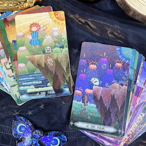 Può includere: Un mazzo di carte dei tarocchi con illustrazioni colorate di paesaggi e figure fantasy. La carta "0 Il Matto" è visibile. Una decorazione a farfalla blu e nera è in primo piano. Una candela accesa e uno specchio con cornice dorata sono sullo sfondo.