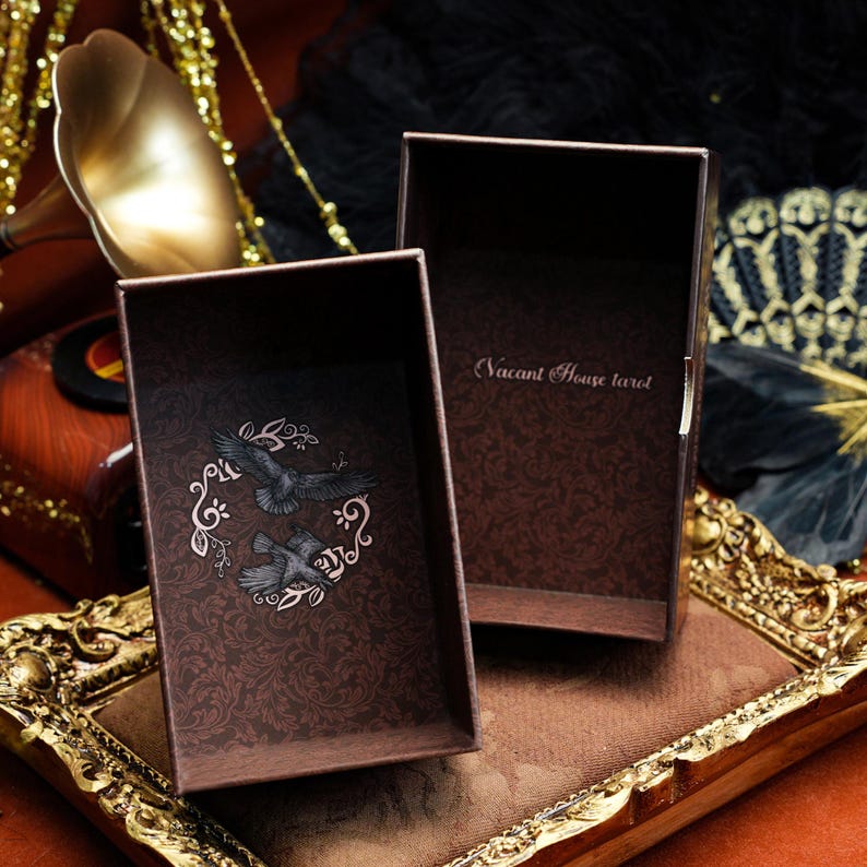 May include: A dark brown rectangular box with an open lid, featuring a repeating damask pattern. The interior base displays two black birds within a circular floral frame, while the inner lid is printed with the text Vacant House tarot.
