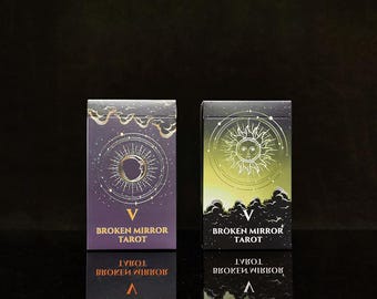 May include: Two rectangular tarot card decks titled Broken Mirror Tarot. The left box is purple with a gold crescent moon and sun design. The right box is green and black with a gold sun illustration. Both feature the Roman numeral V and text.