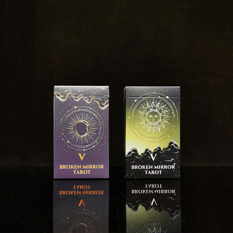 May include: Two rectangular tarot card decks titled Broken Mirror Tarot. The left box is purple with a gold crescent moon and sun design. The right box is green and black with a gold sun illustration. Both feature the Roman numeral V and text.
