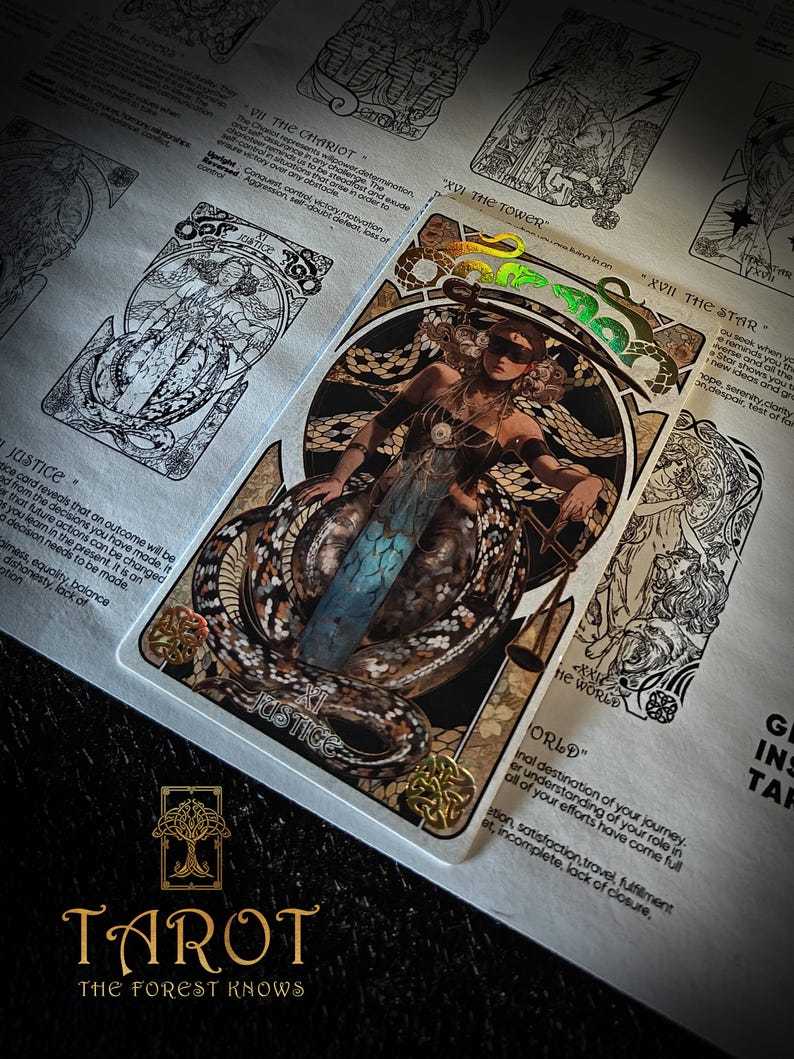 May include: A tarot card depicting a woman with a blindfold and snakes, labelled "JUSTICE". The card is part of a deck with the text "TAROT THE FOREST KNOWS". Other cards are visible in the background, with text describing their meanings.