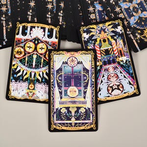 May include: A deck of tarot cards with ornate illustrations. The cards feature celestial and floral designs in a palette of black, gold, and pastel colors. The cards are arranged on a light surface, with some cards fanned out.