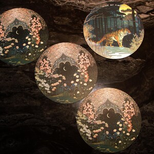 May include: Four round decorative art pieces. Three feature a night scene with a crescent moon, flowers, and an arched structure. The fourth depicts a tiger in a forest with the text "NINE OF FIRE".