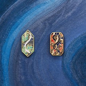 Könnte beinhalten: Zwei Emaille-Pins mit goldenen Metallrahmen. Der erste Pin zeigt eine weiße Kreatur mit blauen und grünen Akzenten auf schwarzem Hintergrund. Der zweite Pin zeigt eine schwarze Kreatur mit roten und blauen Akzenten auf braunem Hintergrund.