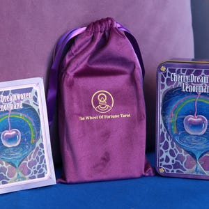 May include: A deck of Cherry Dreamwoven Lenormand cards in a rectangular box, a purple velvet drawstring pouch with a gold logo reading The Wheel Of Fortune Tarot, and a matching rectangular metal tin featuring a cherry and rainbow illustration.