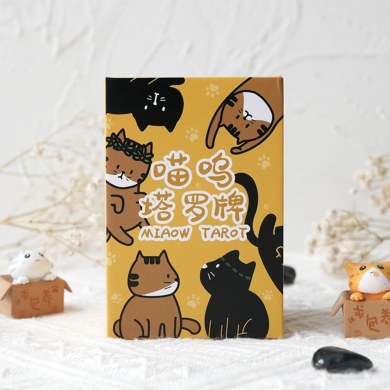 Puede incluir: Una caja amarilla con una baraja de cartas de tarot de gatos. La caja presenta una ilustraci&oacute;n de dibujos animados de gatos sobre un fondo amarillo. El texto en la caja dice "喵喵塔罗 MIAOW TAROT".