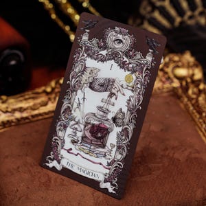May include: A rectangular tarot card titled The Magician featuring a detailed illustration of a hand holding a string attached to a glass bottle containing a dark rose. The card is framed by ornate floral patterns, two black ravens, and an eye.