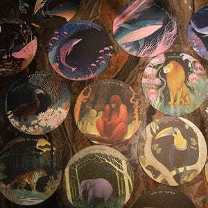 May include: A collection of circular art prints featuring various animals and celestial elements. Each print showcases a unique scene, including a lion, whale, and elephant, with vibrant colors and intricate details. The prints are arranged on a dark, textured surface.