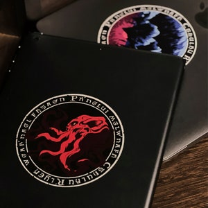 May include: A black laptop with a red and white sticker featuring a circular design with the text "Cenhulu R.Lugher" and "Mglwnaef Phnglui Mglwnaef" in a circular pattern around a red octopus-like creature.