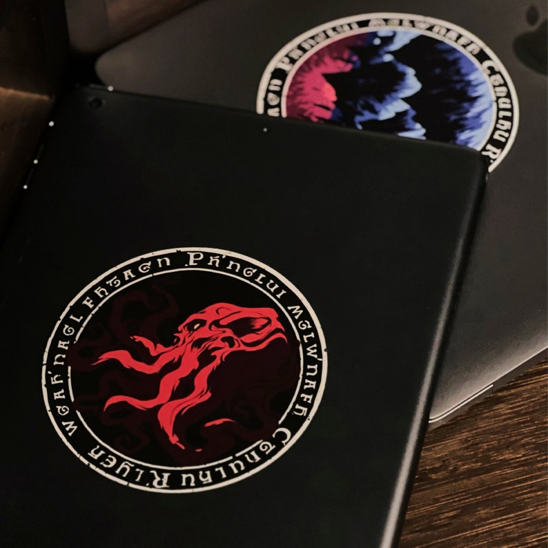 May include: A black laptop with a red and white sticker featuring a circular design with the text "Cenhulu R.Lugher" and "Mglwnaef Phnglui Mglwnaef" in a circular pattern around a red octopus-like creature.